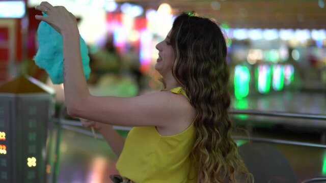 Chica joven con pelo largo tomando algod&oacute;n de az&uacute;car y pas&aacute;ndolo bien en zona de atracciones de una feria