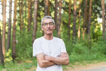 Naklejka na ściany i meble portrait of an elderly man on the background of the forest park. an old man of 60s against the background of trees wearing glasses and a white T-shirt