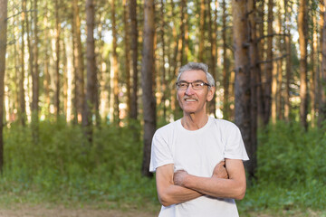 Naklejka na ściany i meble portrait of an elderly man on the background of the forest park. an old man of 60s against the background of trees wearing glasses and a white T-shirt. the concept of a happy and prosperous retirement