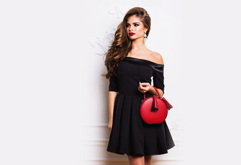 Amazing luxury seductive woman in stylish black party dress posing on white wall . Red hand bag ....