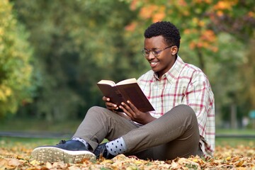 Portrait of happy positive black African man, Afro American ethnic intelligent young guy, reader in glasses, shirt is smiling walking outdoors in golden autumn park, sitting on ground, reading book