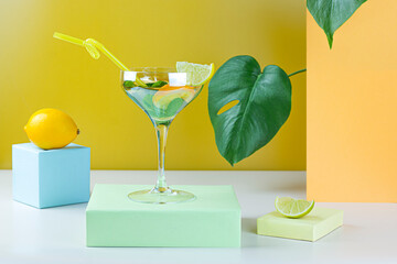 Cocktail in vintage champagne glass with mint leaves, lime and lemon on pastel color podiums. Summer cocktail with Monstera leaves on yellow and white background. Abstract concept. Part of set.