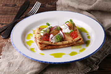 Russian and Ukrainian cuisine. Pancakes with sour cream, caviar and herbs on a white plate on a wooden background. Rustic. Burlap, fork and knife. Traditional dish. Background image.