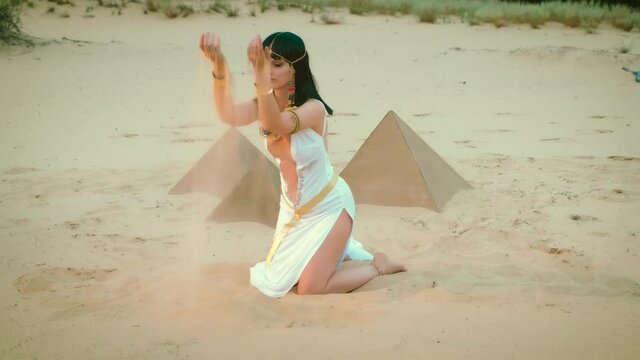Beautiful sexy charming woman in image Egyptian pharaoh takes hands yellow sand, plays, conjures pours out. Hairstyle girl brunette princess decorated gold chain, necklace, bracelets. Pyramids, desert