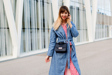 Happy smiling woman in fashionable blue coat and striped red dress walking over modern business...