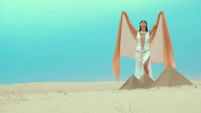 Egyptian beautiful woman goddess prays, raises hands up on backdrop pyramids, yellow sand, blue azure watercolor clear sky. Orange cape scarf, white sexy luxury retro vintage dress. Brunette long hair