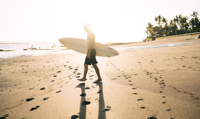Surf guy go to summer surfing in summer ocean for catching waves during morning sunshine, young man holding professional blank surfboard with mockup copy space area visiting Bali island, Indonesia