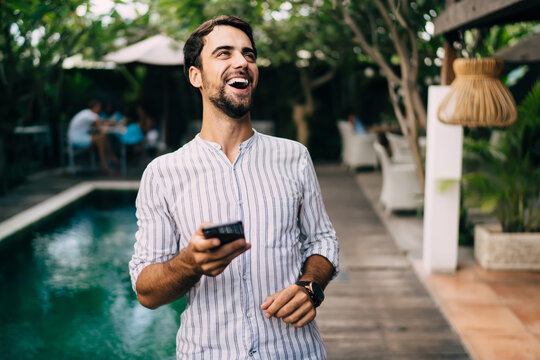 Cheerful guy laughing happily while browsing smartphone standing in tropical garden
