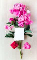 on a light wooden background there is a bouquet of pink peonies, and on top is a white card with a place for text, next to it are hearts, rose and peony petals, a red box with a gift. copy space