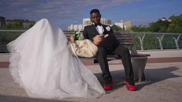 Wide shot of smiling happy groom sitting on bench with bride lying on knees. Romantic African American couple of newlyweds talking on wedding day enjoying nature outdoors. Romance concept