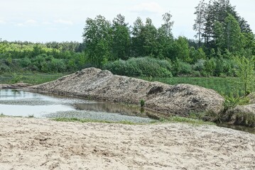 part of the lake with heaps of gray sand on the shore against the background of green vegetation and the sky