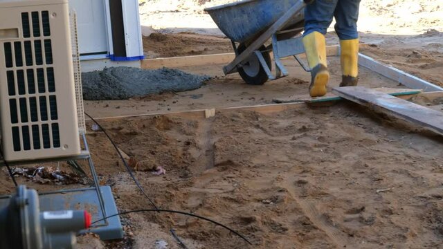 Construction worker in yellow boots pushing wheelbarrow with concrete upa plank at house building site, then he dumps it in the wooden form for a patio.