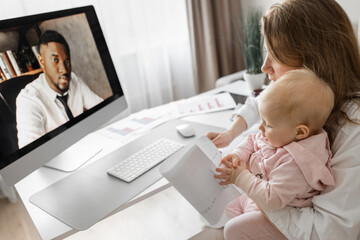 Carrying mother in maternity leave engaged in remote work or study at home on PC while infant baby girl hold documents and looking at screen. Modern day mother of little kid sit at desk use gadgets