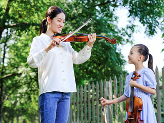 The female teacher instructs the girl to play the violin in the outdoor park