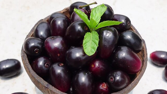 Dark pink-red ripe Syzygium cumini fruits. Dark black java plum in a wood bowl at isolated white background. Green mint leaf on top of some large java plums. 4k video.