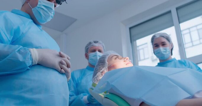 Male and female medical workers in disposable masks, hats are standing near the dental chair with a female patient with an anesthesia tube who is going up from the lay position to sit.