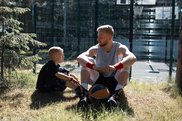 Sportive young father and son basketball players sitting on lawn with ball. Happy parenting concept