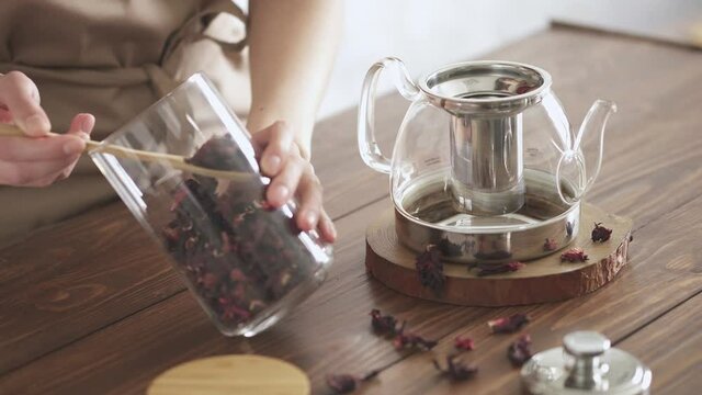 Red tea brewing process. Step one. A young housekeeper sprinkles dry hibiscus tea from a glass jar with a spoon. Pour hibiscus tea into a glass teapot. Making red herbal tea on a wooden kitchen table.