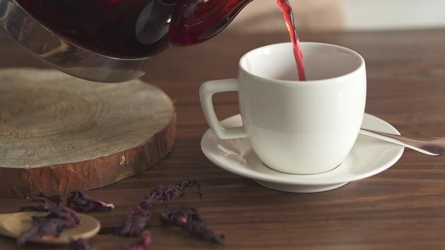 Hot red tea pouring into a mug close-up. Step four. The process of pouring red tea into a white cup. Glass teapot with hot tea on a wooden cut. In the foreground is dry hibiscus tea in a wooden spoon
