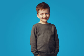Kid smiling while holding hands in pockets, isolated over blue background