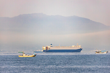 Large ship and small fishing boats sail by misty mountain