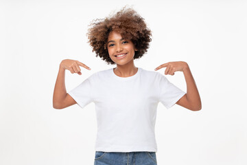 Smiling african girl pointing at her blank white t-shirt with both index fingers, copy space for...