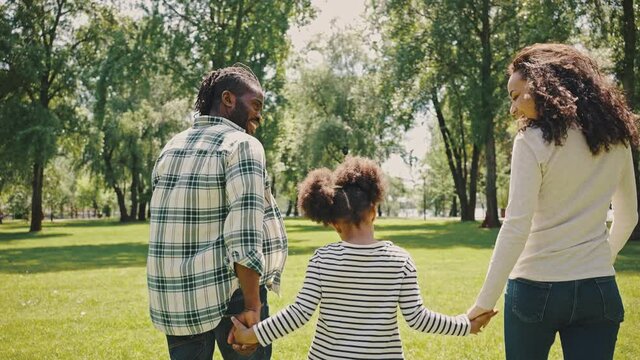 Rear view of Happy mixed race family parents with cute child girl holding hands and walking together in the park. Father and mother with daughter enjoy and having fun in summer outdoor vacation