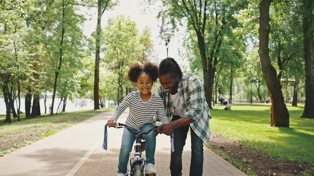 Close up portrait of father teaches his daughter to ride a two-wheeled bike. Childrens bike, a girl learns to ride for the first time. Happy Holidays African Ethnicity Family