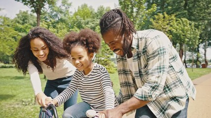 African American parents teaching their little girl to driving bike in park. Happy family with excited daughter learning to ride a bike for the first time - Powered by Adobe
