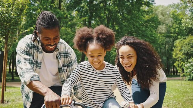 Happy family with excited daughter learning to ride a bike for the first time. African American parents teaching their little girl to driving bike in park