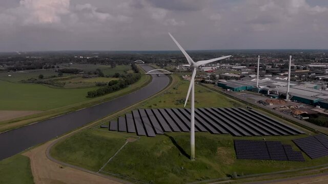 Aerial approach of a rotating wind turbine and solar panels in The Netherlands part of sustainable industry in Dutch flat river landscape against blue sky with clouds. Circular economy concept.