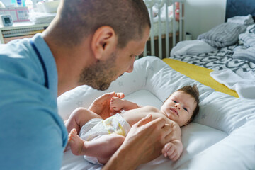 Obraz premium Caucasian man father taking care of his two months old baby boy or girl on the bed at home changing diapers and clothes - father's day parenthood concept real people family