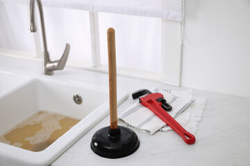 Plunger, pipe wrench and towel on kitchen counter near clogged sink