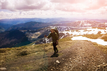 Obraz premium Young smiling girl climbs the mountains with trekking poles and a backpack in early spring, on the background there is a beautiful view with snowy mountains. Traveler goes hiking in the national park.