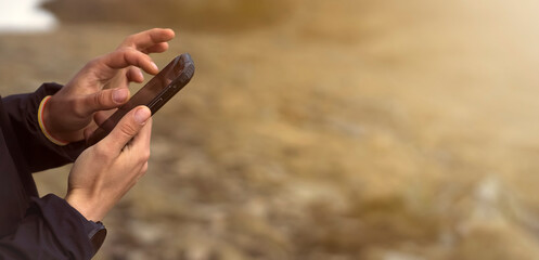 Man holds a smartphone in his hands and looks at a map, makes a route through the mountains, writes a message, chats. Young traveler explores the area, uses the app on his device while hiking.