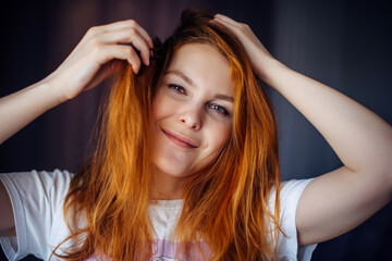 Fototapeta premium Portrait of beautiful young woman with thick, tousled red hair, close-up. Green-eyed beauty touches her hair while looking at camera and smiling. Advertising of care, hair coloring.