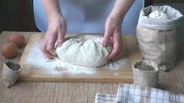 The hands of an elderly woman knead the dough for bread on a wooden board on the table, on the table there are 2 eggs, there are linen bags with spices cumin and coriander and flour. World Bread Day