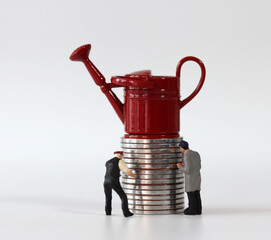 Red miniature watering can on a pile of coins and two miniature people. Miniature people and coin...