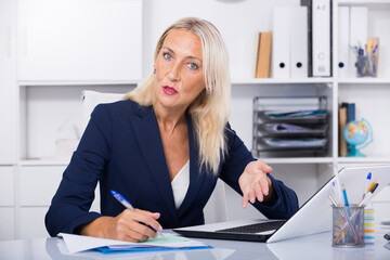Focused mature female entrepreneur sitting at office with papers and laptop
