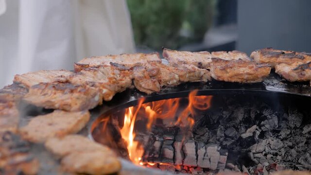 Slow motion: process of cooking fresh juicy meat steaks on round brazier with hot flame at summer local food market - close up view. Outdoor cooking, gastronomy, cookery, street food concept