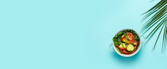 Vegetable salad in a blue bowl and a green leaf of a palm tree on a blue background. Top view, flat lay. Banner
