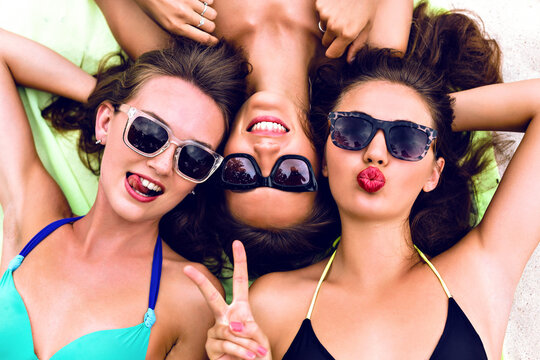 Close up lifestyle summer portrait of thee girls friends relaxed and getting sunbathe, laying on the beach, wearing bright bikinis and stylish vintage sunglasses. Swing tongue, sending kiss.