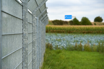 Fototapeta na wymiar Gray steel fence and agricultural vegetable fields. blue town sign. Selective focus.