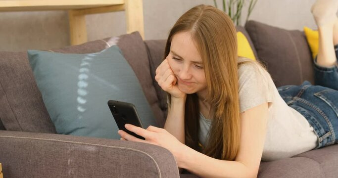 Disgruntled upset girl scrolls through the news feed on Internet. Woman lies on the couch with smartphone