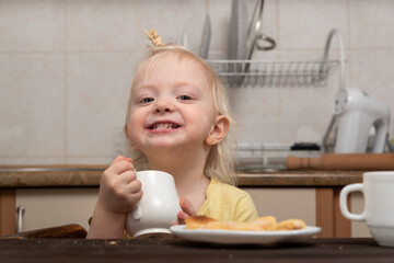 Little smiling girl drinks a milk. Breakfast with child. Cute blonde child with cup in hands in kitchen.