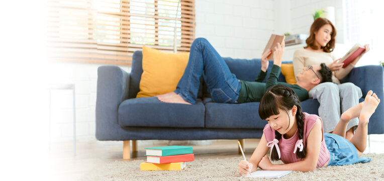 Relax Asian parent reading a book on sofa and daughter painting art in living room at home. panoramic background..