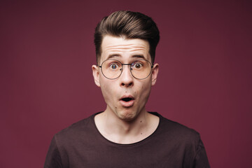 Surprised male, keeps mouth opened, being stupefied as advertises something, wears glasses, brown t shirt over crimson background. Omg concept