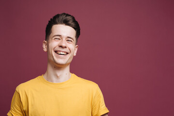 Isolated shot of young laughing handsome male with trendy hairdo, wears casual brown t shirt, has serious expression as listens to interlocutor, poses in studio against crimson background