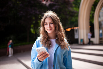 Fototapeta premium Young woman on Wednesday street with a phone in her hands. A beautiful, happy girl looks at the camera and listens to music on headphones. 