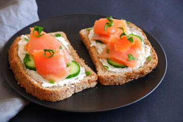 Toast with red fish, curd cheese and cucumber on a black plate.